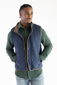 A man wearing a navy blue quilted vest with tan trim over a green button-up shirt and blue jeans, photographed against a plain white studio background.