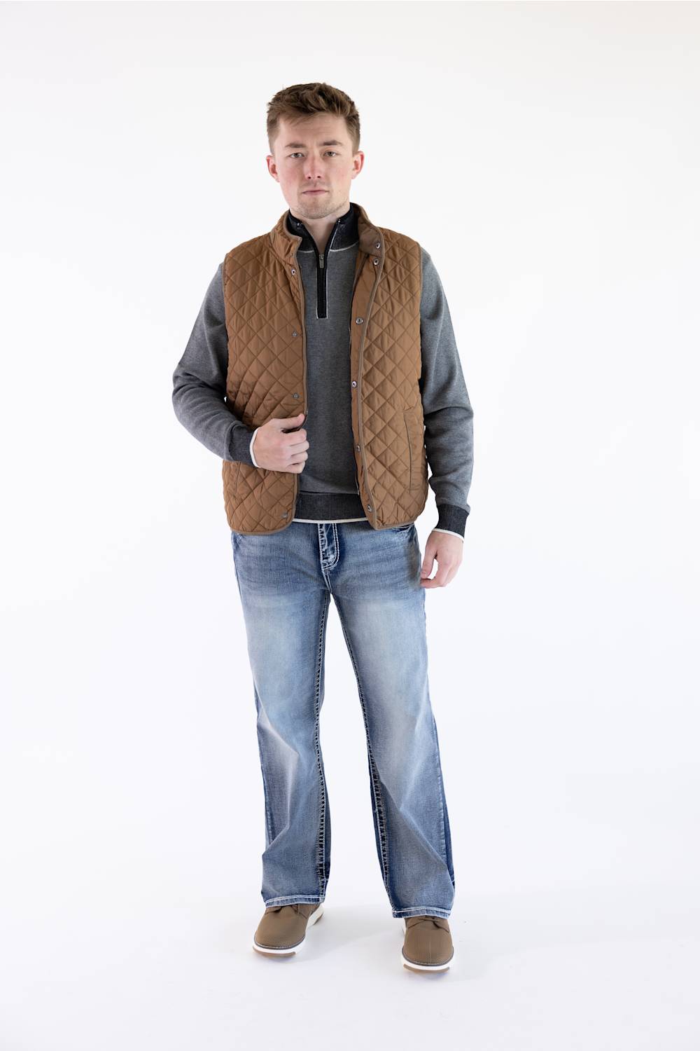 Full-length view of a model wearing a brown rhombus quilted vest, gray mock-neck sweater, medium-wash jeans, and brown shoes.