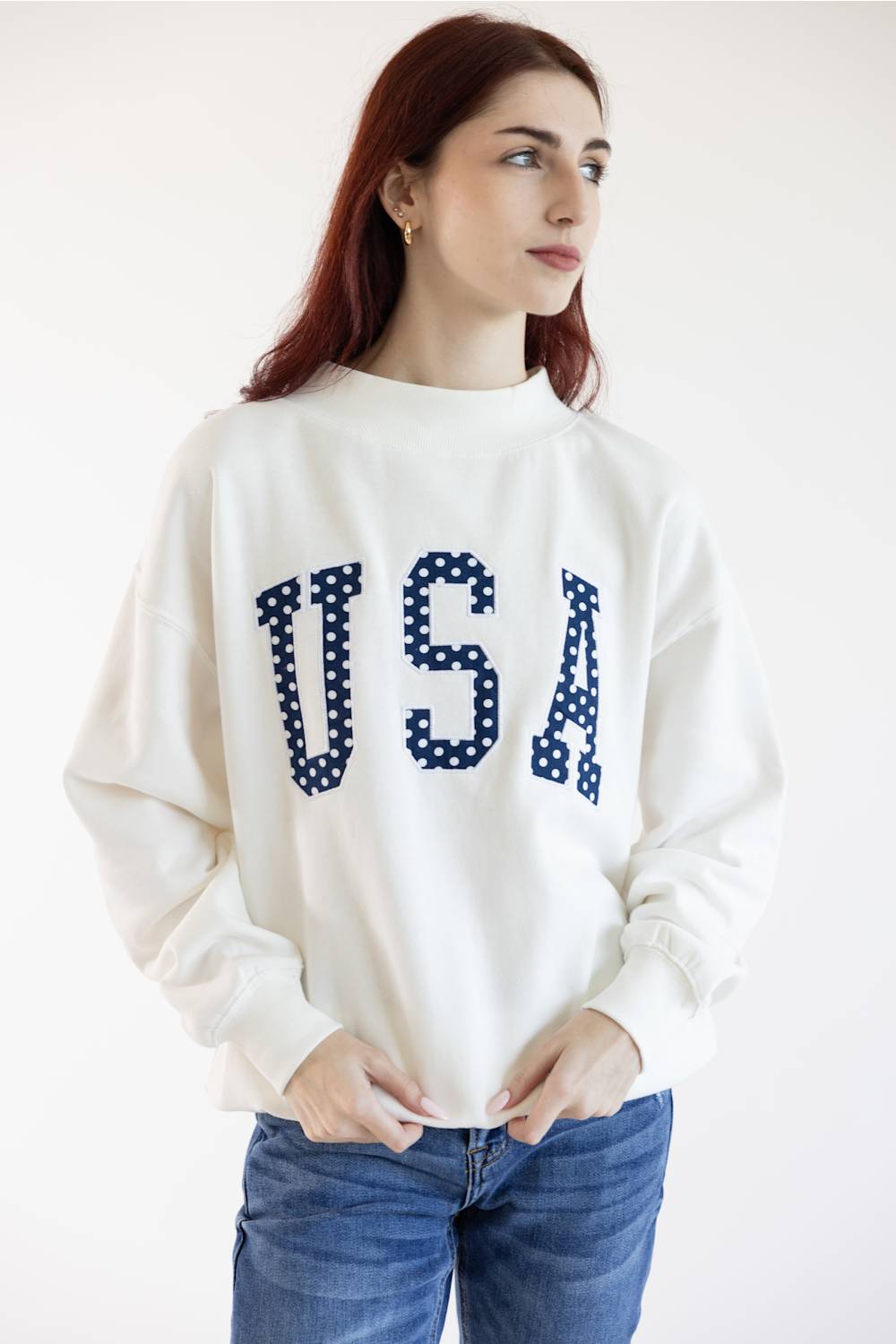 A woman with red hair wearing a white crewneck sweatshirt with 'USA' written across the chest in navy blue letters filled with white polka dots, paired with blue jeans, against a plain white background.