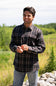 A smiling male model standing outdoors in a sunny field, wearing the Split Flannel Shirt for Men in Slate with a charcoal gray, burgundy, and cream plaid pattern, dual buttoned chest pockets, and light wash jeans.
