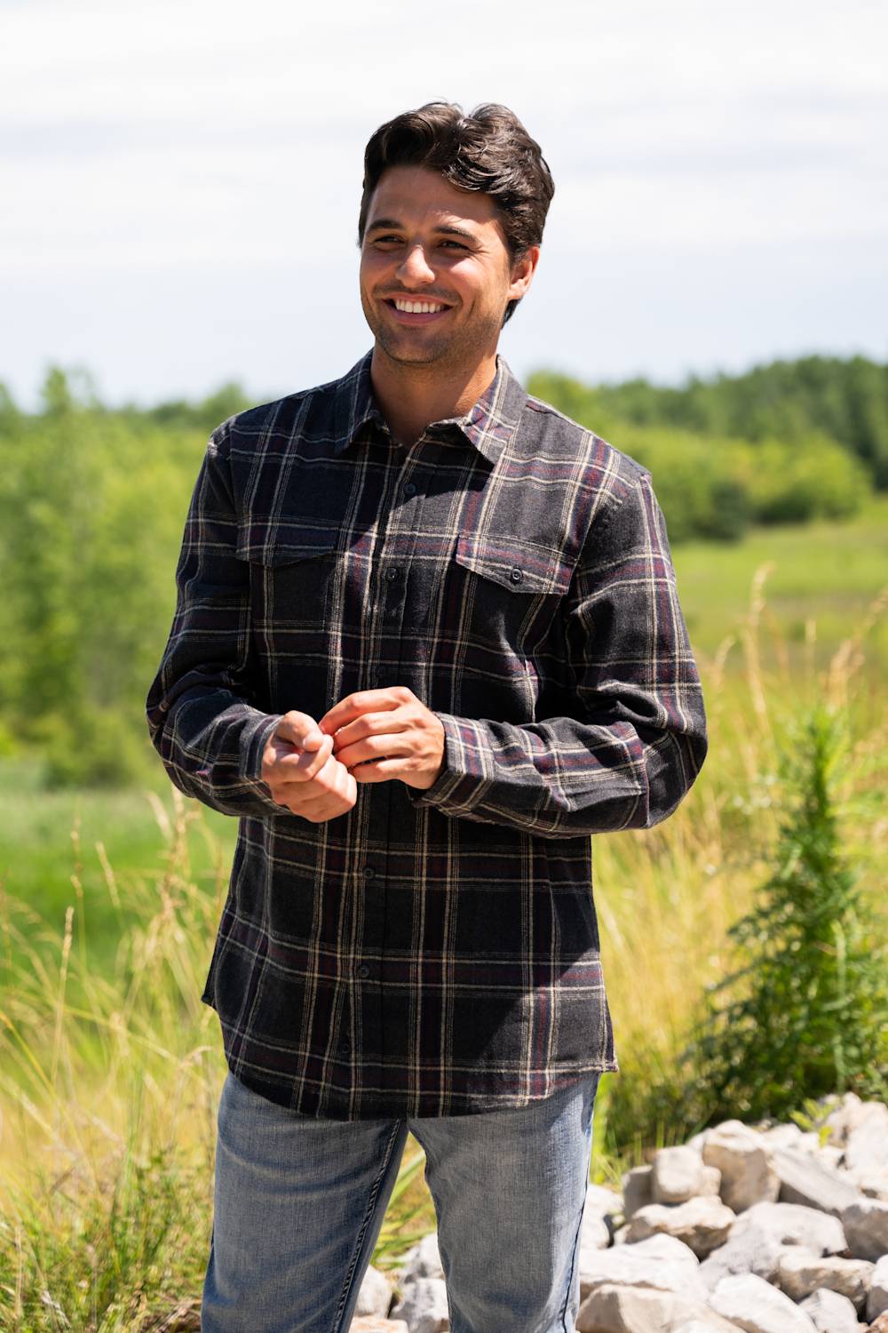 A smiling male model standing outdoors in a sunny field, wearing the Split Flannel Shirt for Men in Slate with a charcoal gray, burgundy, and cream plaid pattern, dual buttoned chest pockets, and light wash jeans.