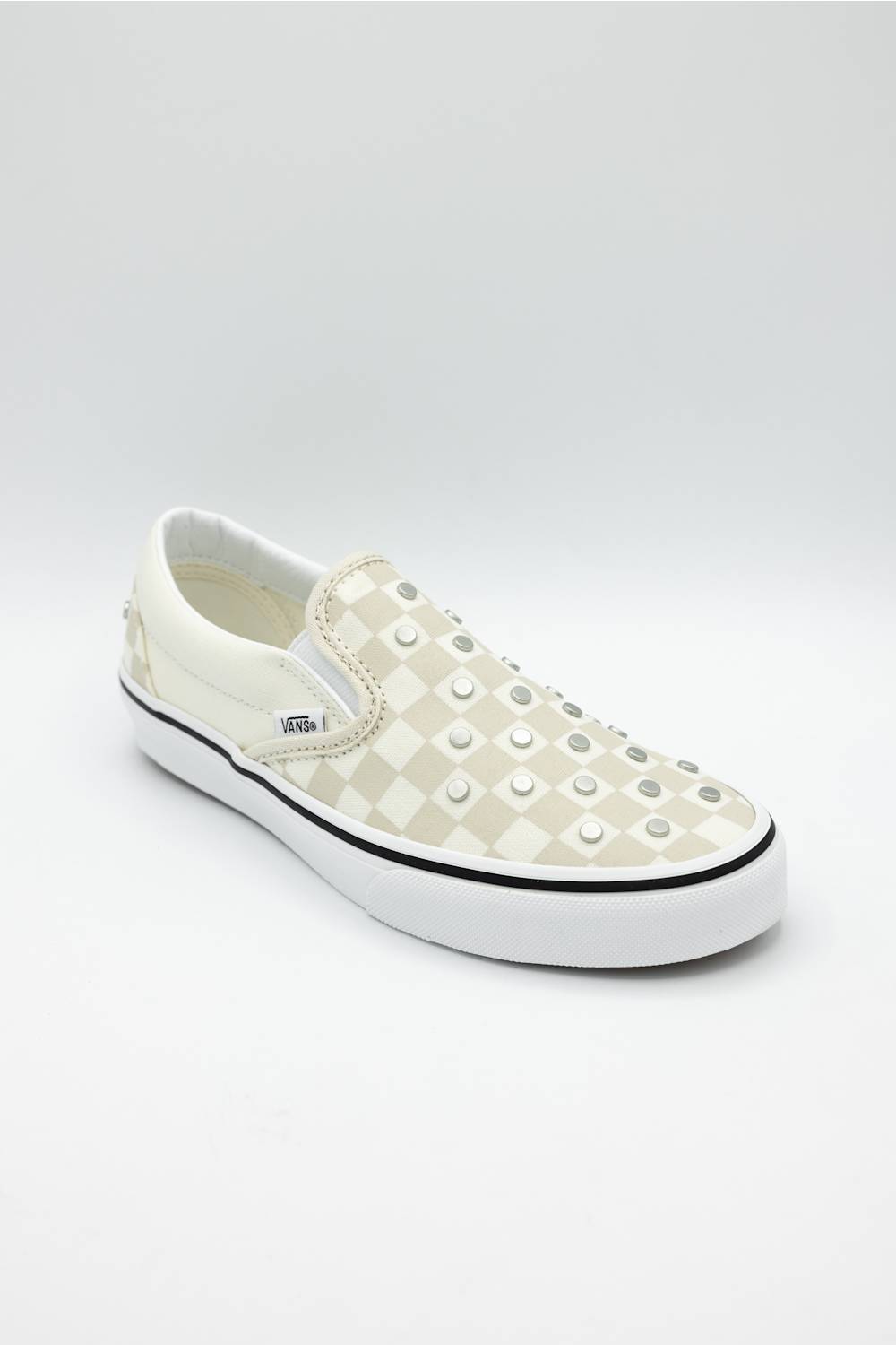 A white slip-on platform sneaker featuring a checkerboard pattern on the side and rows of silver metal studs across the toe box, shown against a light gray studio background.