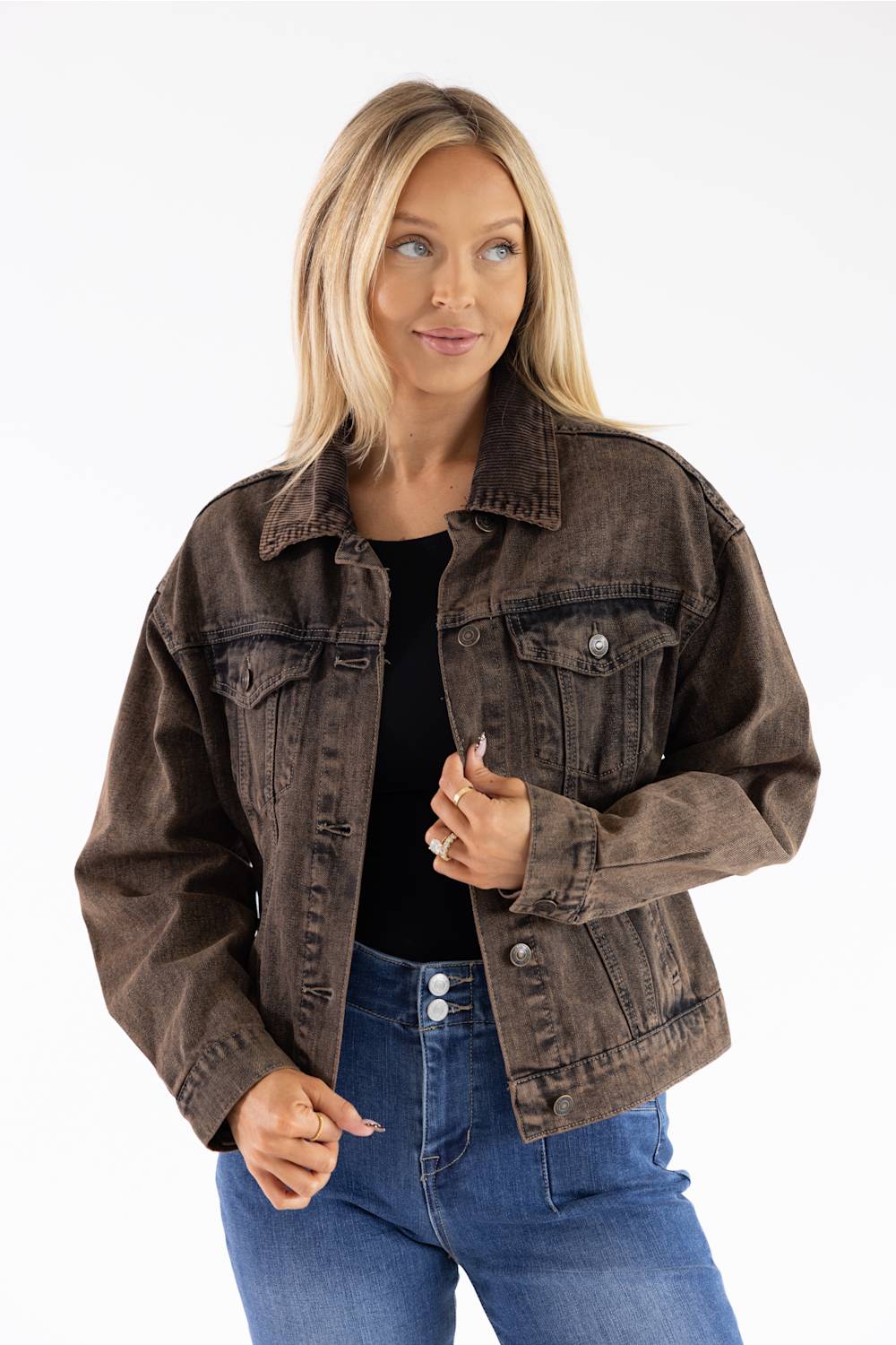 A woman modeling a dark brown washed denim jacket over a black top and blue jeans, photographed against a plain white studio background.