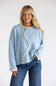 A woman modeling a light blue fuzzy knit sweater with a cream zigzag trim and button detail down the front, paired with blue jeans, against a plain white studio background.