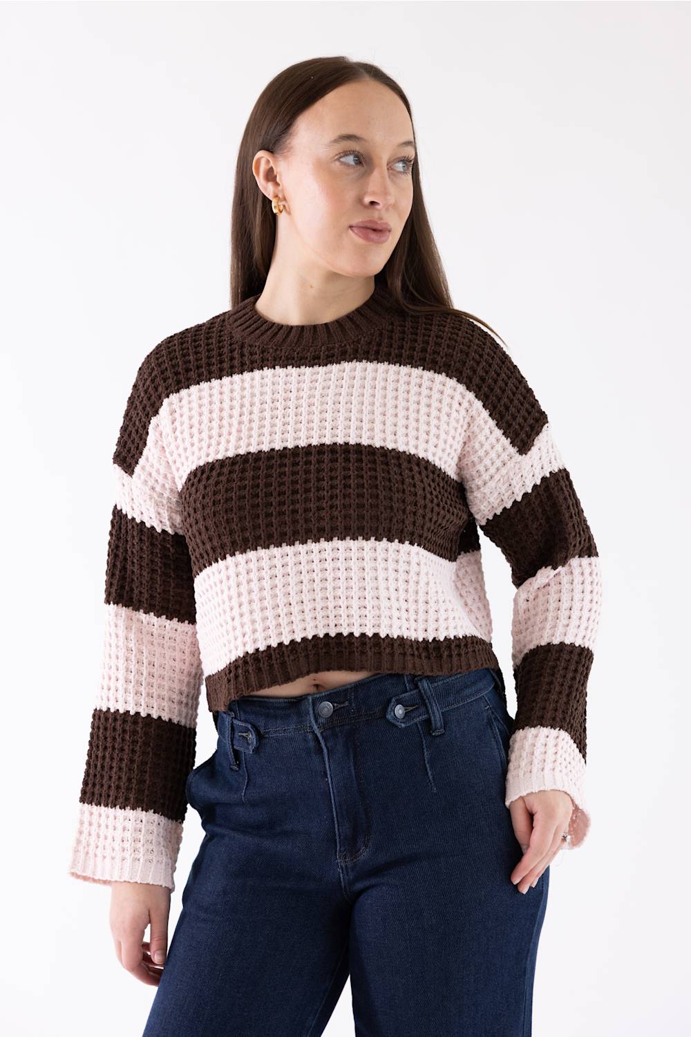 A woman wearing a brown and light pink wide-striped knit crew neck sweater with dark blue jeans, photographed against a plain white studio background.