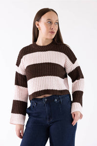 A woman wearing a brown and light pink wide-striped knit crew neck sweater with dark blue jeans, photographed against a plain white studio background.