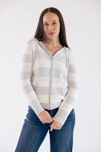 A woman wearing a cream and light gray striped zip-up hoodie with a hood and front pockets, paired with blue jeans, standing against a plain white studio background.