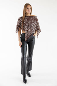 A full-length fashion photograph of a woman wearing a brown floral scarf poncho top, black faux-leather flare pants, and black boots against a white studio background.