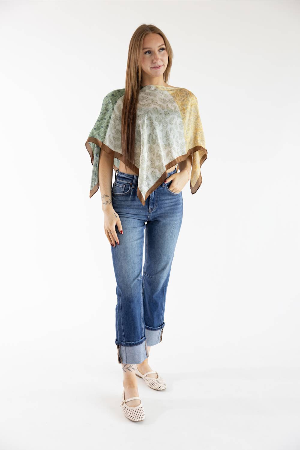 A woman modeling a lightweight poncho-style top with a green, beige, and brown abstract pattern, paired with blue jeans and white pointed flats, against a plain white studio background.