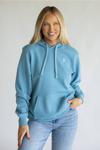 A woman wearing a light blue pullover hoodie with a small white embroidered logo on the left chest, paired with light-wash blue jeans, standing against a plain light gray background.