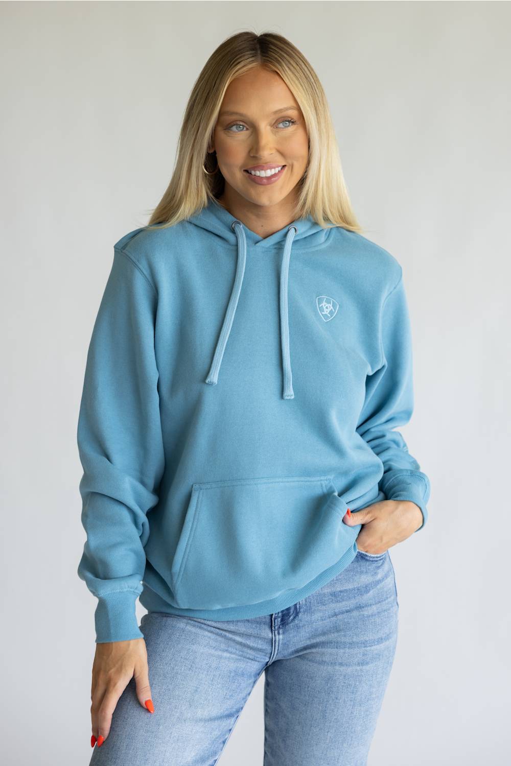 A woman wearing a light blue pullover hoodie with a small white embroidered logo on the left chest, paired with light-wash blue jeans, standing against a plain light gray background.