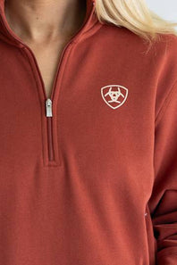 A woman modeling a rust-colored quarter-zip sweatshirt with a small shield logo on the left chest, paired with light blue jeans, against a light gray studio background.