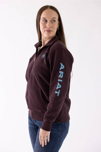 A woman modeling a dark maroon quarter-zip pullover sweatshirt with a light blue vertical 'ARIAT' logo on the sleeve and a matching logo patch on the chest, paired with blue jeans against a white studio background.