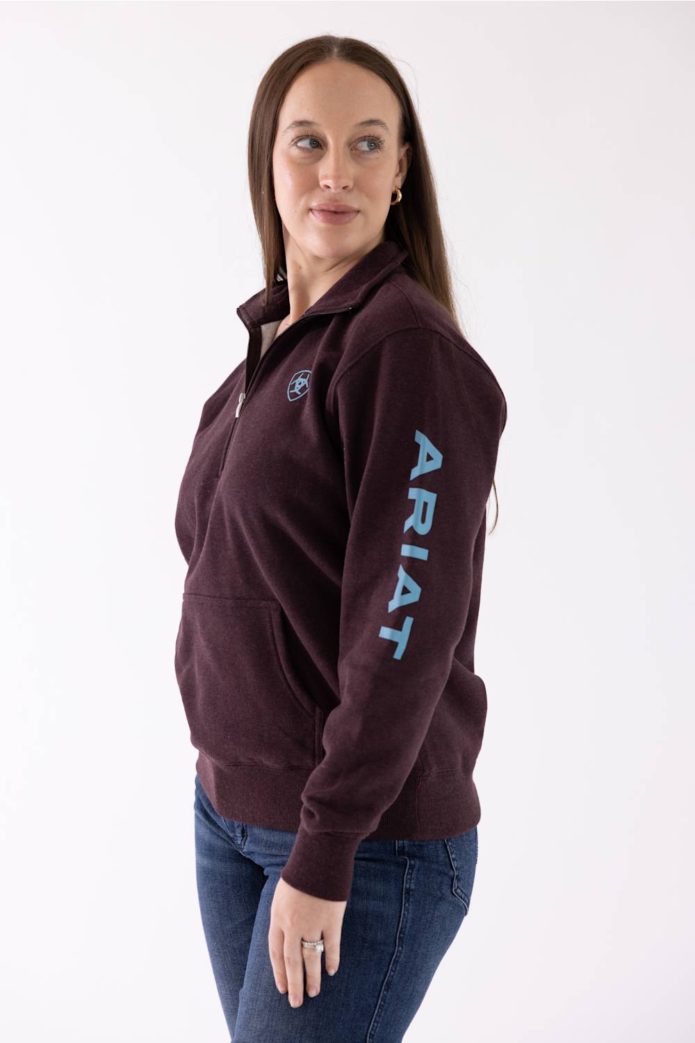 A woman modeling a dark maroon quarter-zip pullover sweatshirt with a light blue vertical 'ARIAT' logo on the sleeve and a matching logo patch on the chest, paired with blue jeans against a white studio background.