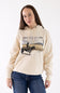 Front view of a model wearing the parchment beige Ariat Wish You Were Here Hoodie, featuring a western graphic of a cowboy on horseback, a front kangaroo pocket, and drawstrings, paired with dark blue jeans.
