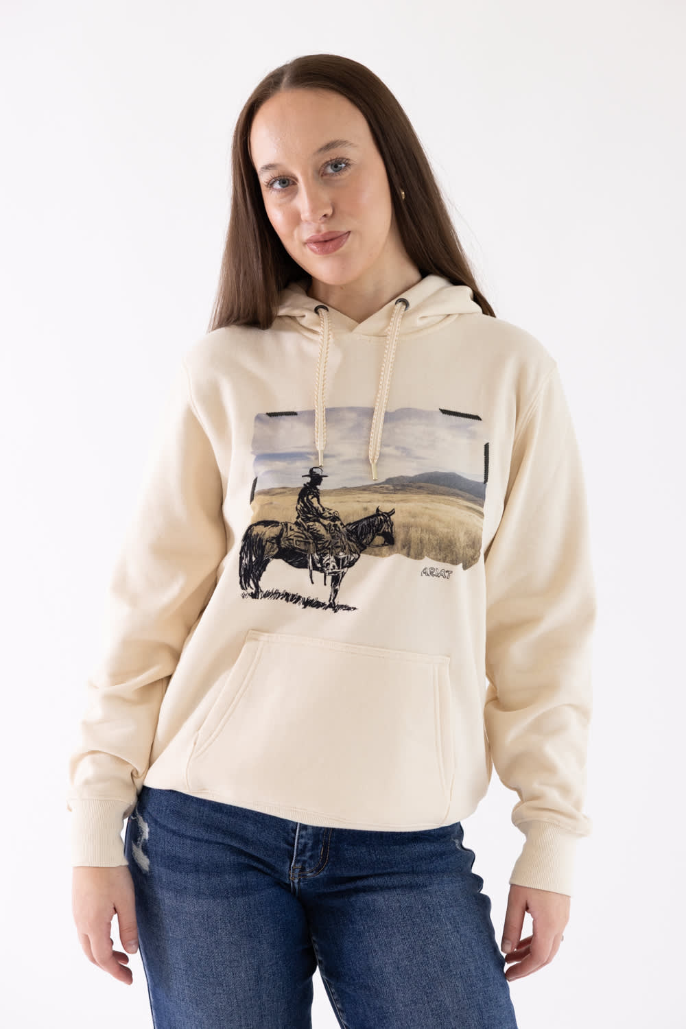 Front view of a model wearing the parchment beige Ariat Wish You Were Here Hoodie, featuring a western graphic of a cowboy on horseback, a front kangaroo pocket, and drawstrings, paired with dark blue jeans.