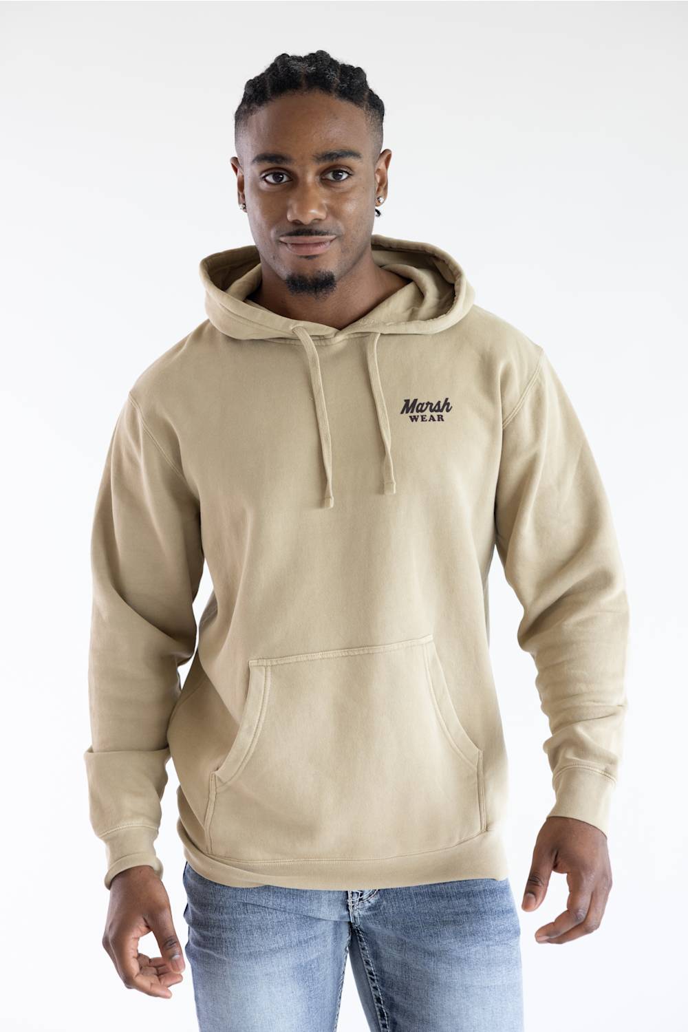 A man wearing a beige pullover hoodie with a drawstring hood and kangaroo pocket, featuring a small black embroidered 'Marsh Wear' logo on the left chest, paired with blue jeans against a plain white studio background.