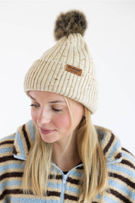 A woman wearing a cream ribbed knit pom-pom beanie with a small brown brand patch and a blue, brown, and cream striped fleece zip-up sweater, photographed against a clean white background.