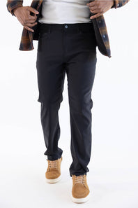 A person wearing slim-fit black trousers, styled with a white t-shirt, an open brown plaid flannel shirt, and tan suede sneakers, shown from the waist down on a white studio background.