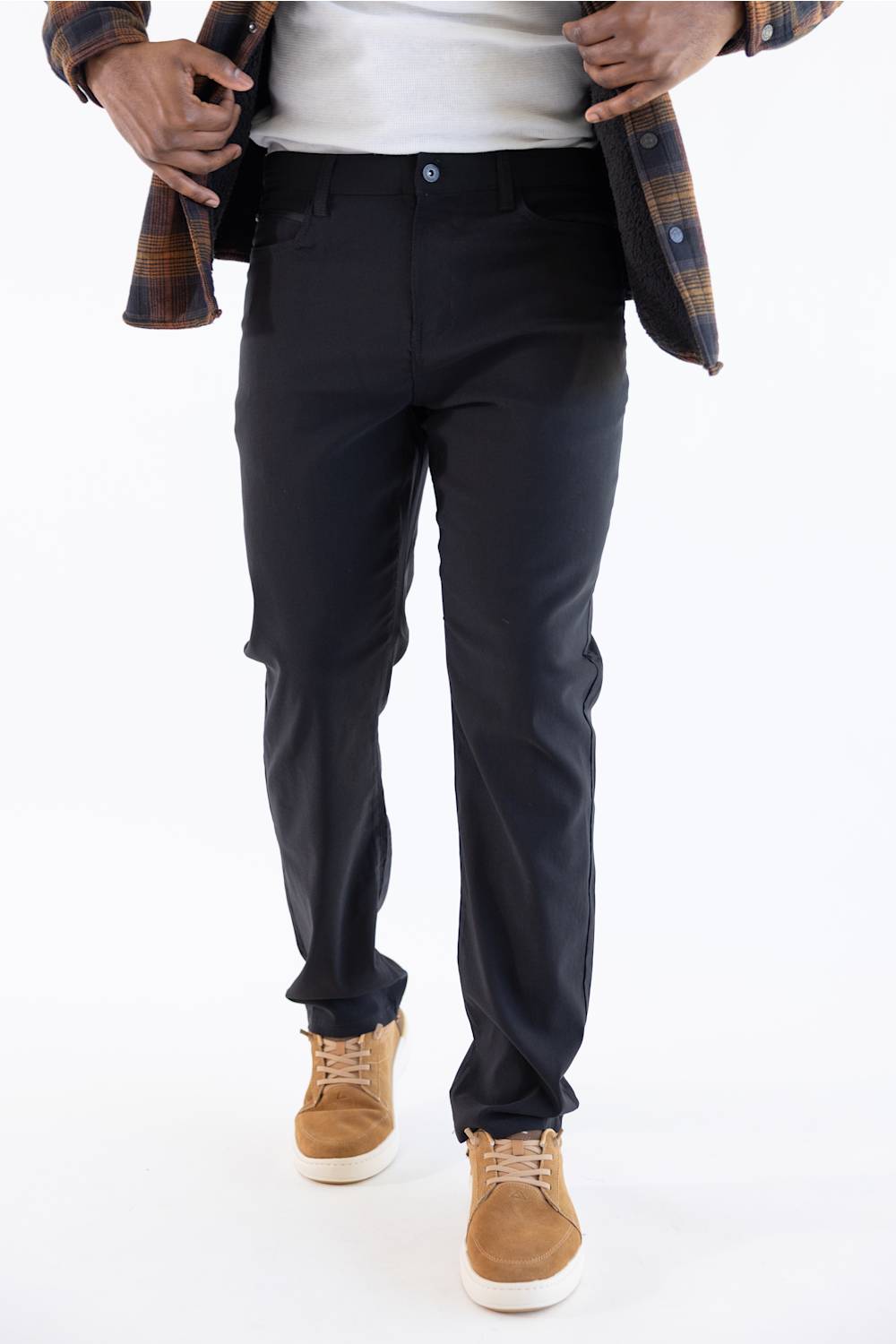 A person wearing slim-fit black trousers, styled with a white t-shirt, an open brown plaid flannel shirt, and tan suede sneakers, shown from the waist down on a white studio background.