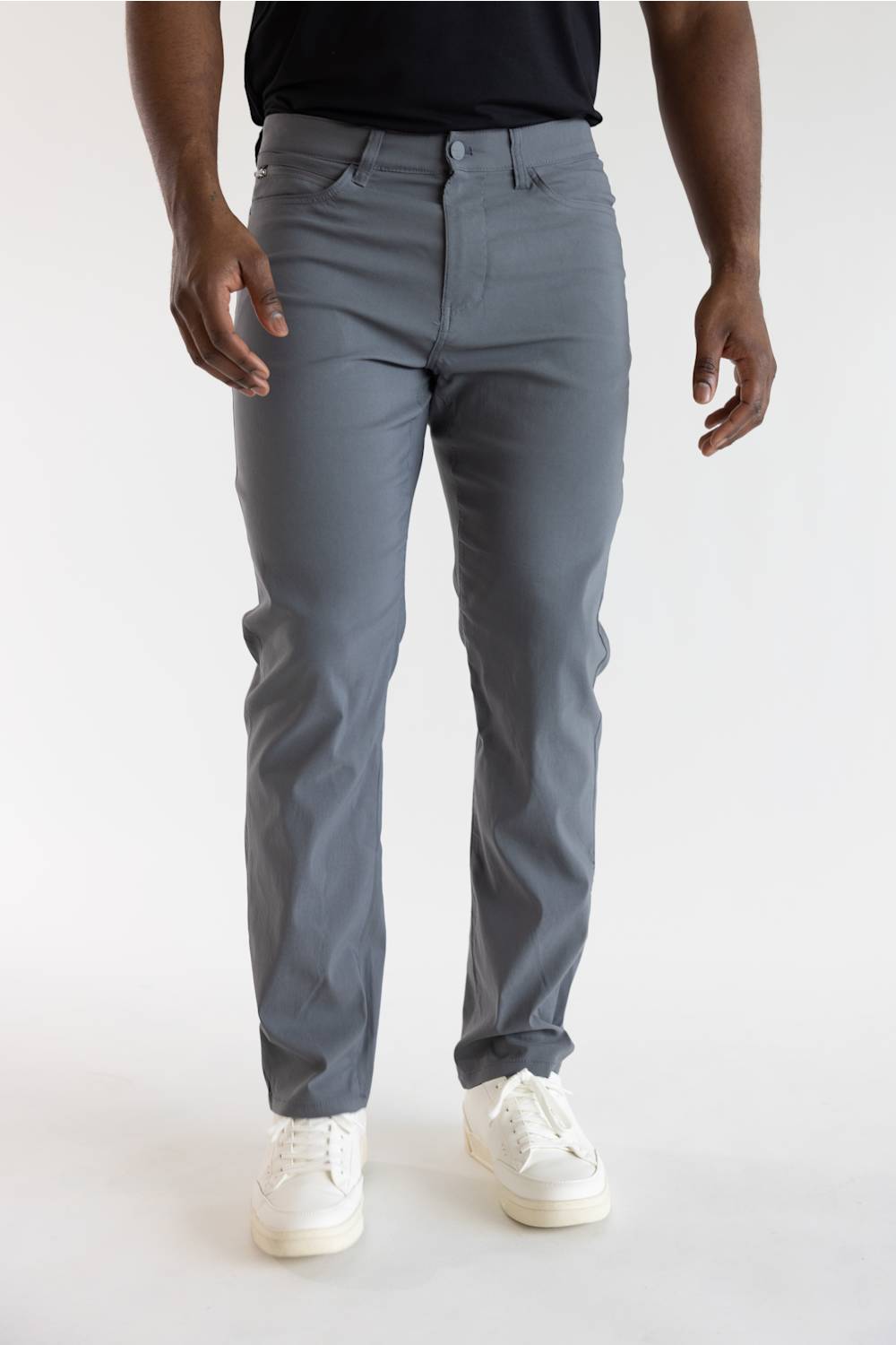 A person wearing slim-fit gray trousers, paired with a black top and white sneakers, standing against a plain light gray studio background.