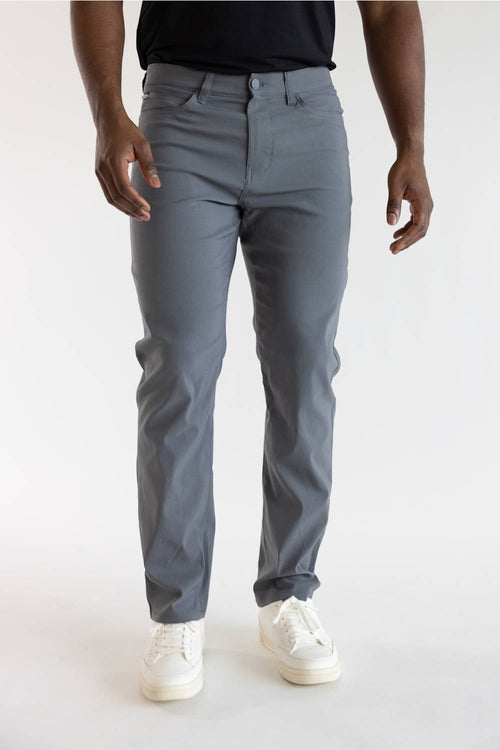 A person wearing slim-fit gray trousers, paired with a black top and white sneakers, standing against a plain light gray studio background.