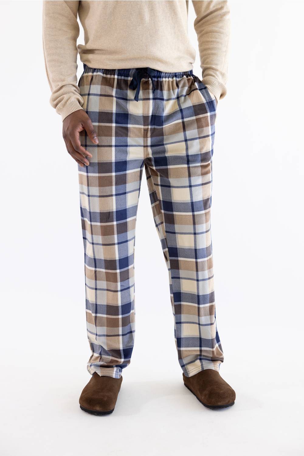 A person wearing plaid lounge pants in beige, blue, and brown, paired with a cream sweater and brown slippers, shown from the waist down on a white studio background.