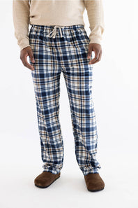 A person wearing blue, white, and beige plaid lounge pants with a drawstring waist, paired with a cream sweater and brown slippers, photographed against a plain white studio background.