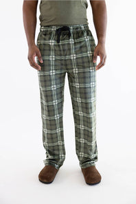 A person wearing green and white plaid pajama pants with a dark drawstring, paired with a muted green shirt and brown slippers, shown against a plain white studio background.