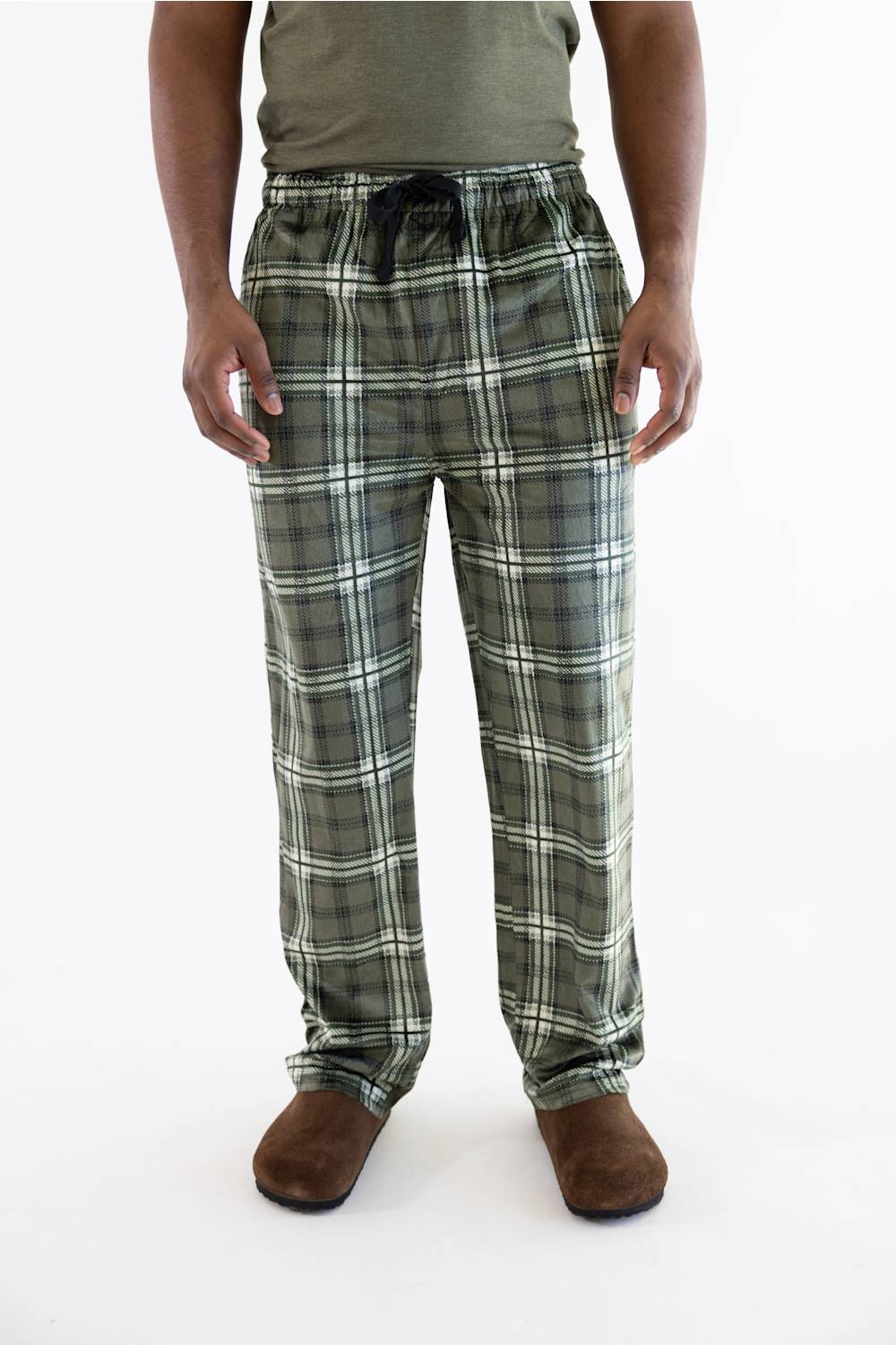 A person wearing green and white plaid pajama pants with a dark drawstring, paired with a muted green shirt and brown slippers, shown against a plain white studio background.