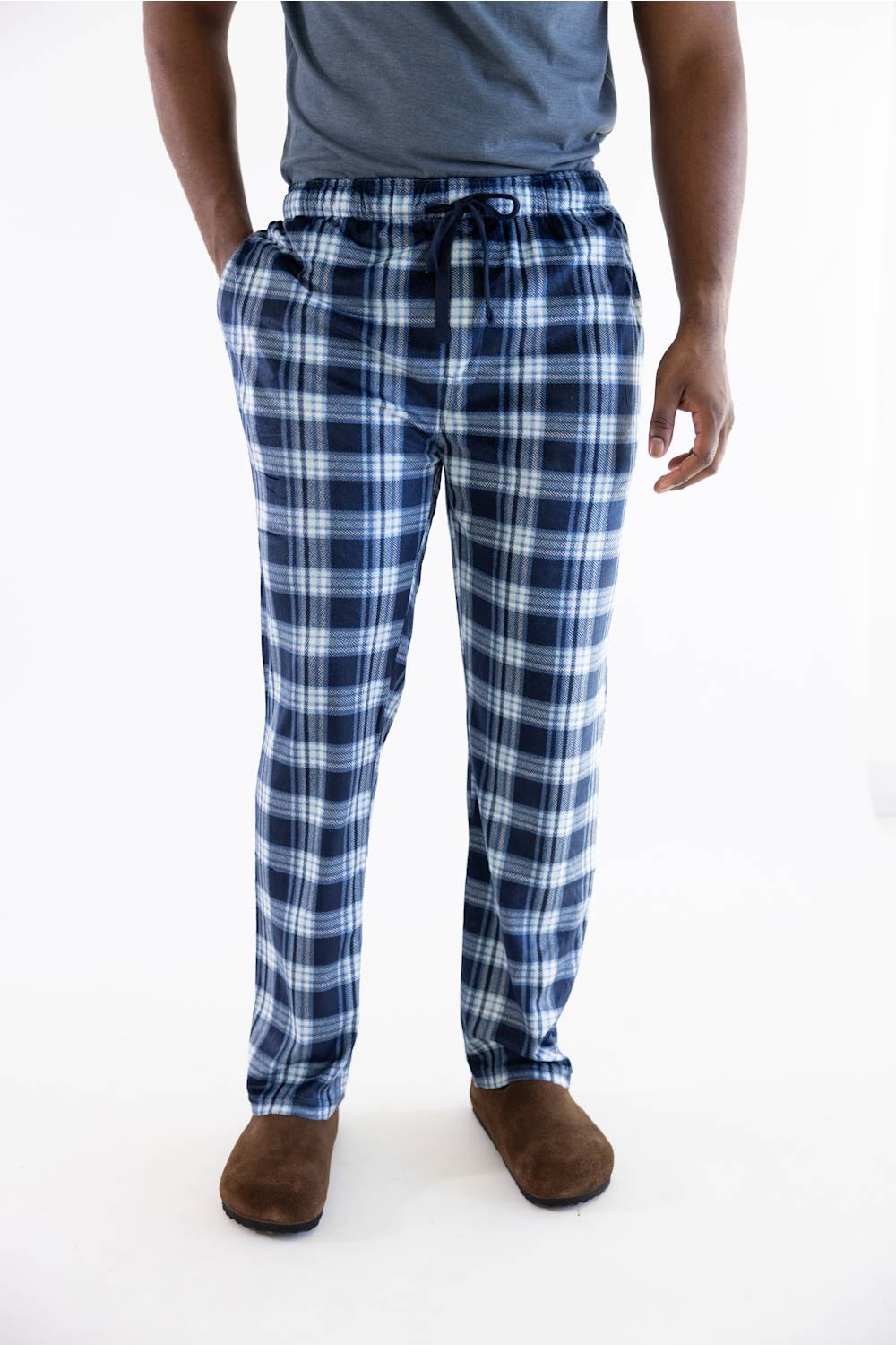 A person wearing blue and white plaid pajama pants with a drawstring waist, paired with a gray t-shirt and brown slippers, shown from the waist down on a white studio background.