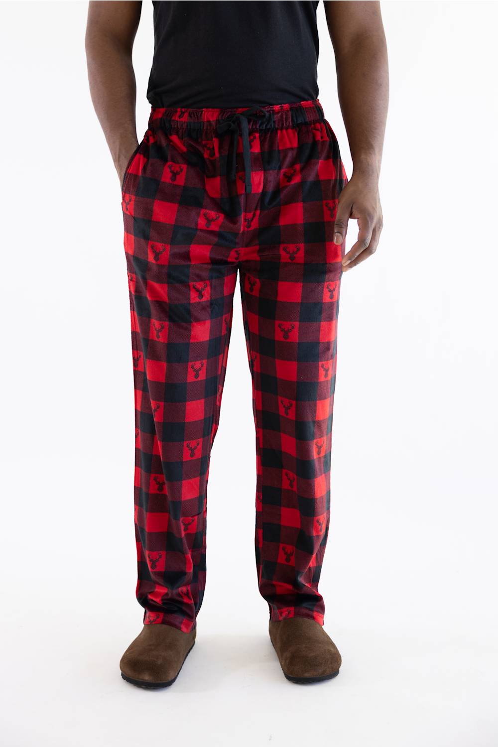 A person wearing red and black buffalo plaid pajama pants with a drawstring waist, paired with a black shirt and brown slippers, shown from the waist down on a white studio background.