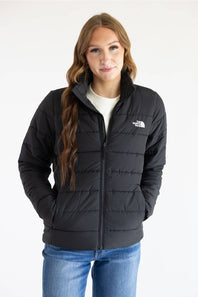 A woman wearing a black puffer jacket with a white embroidered logo on the left chest, paired with a white shirt and blue jeans, against a plain white background.