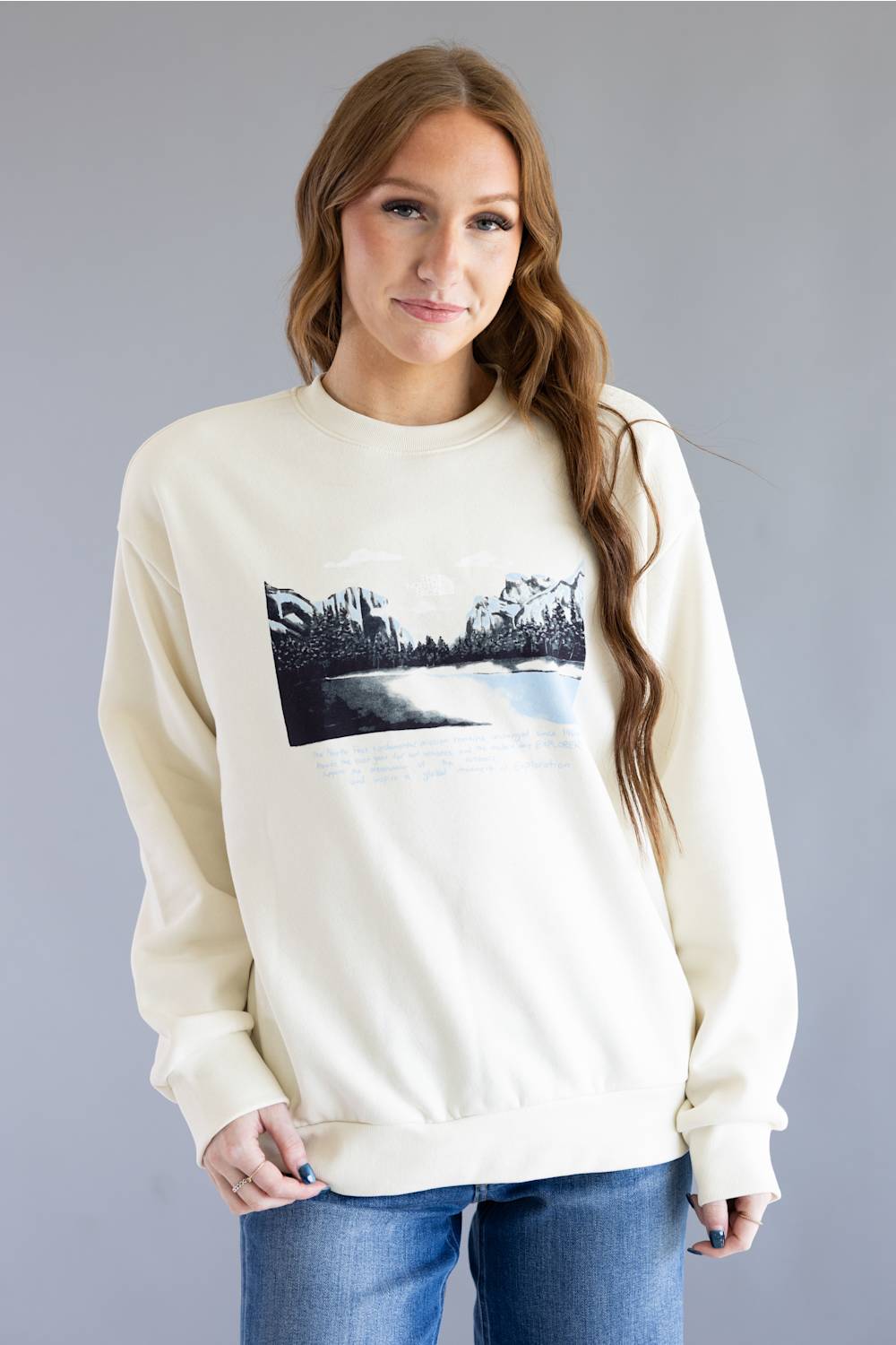 A woman wearing a cream-colored crewneck sweatshirt with a black and white landscape graphic on the front, paired with blue jeans, set against a solid gray studio background.