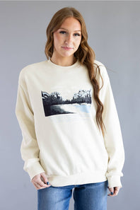 A woman wearing a cream-colored crewneck sweatshirt with a black and white landscape graphic on the front, paired with blue jeans, set against a solid gray studio background.