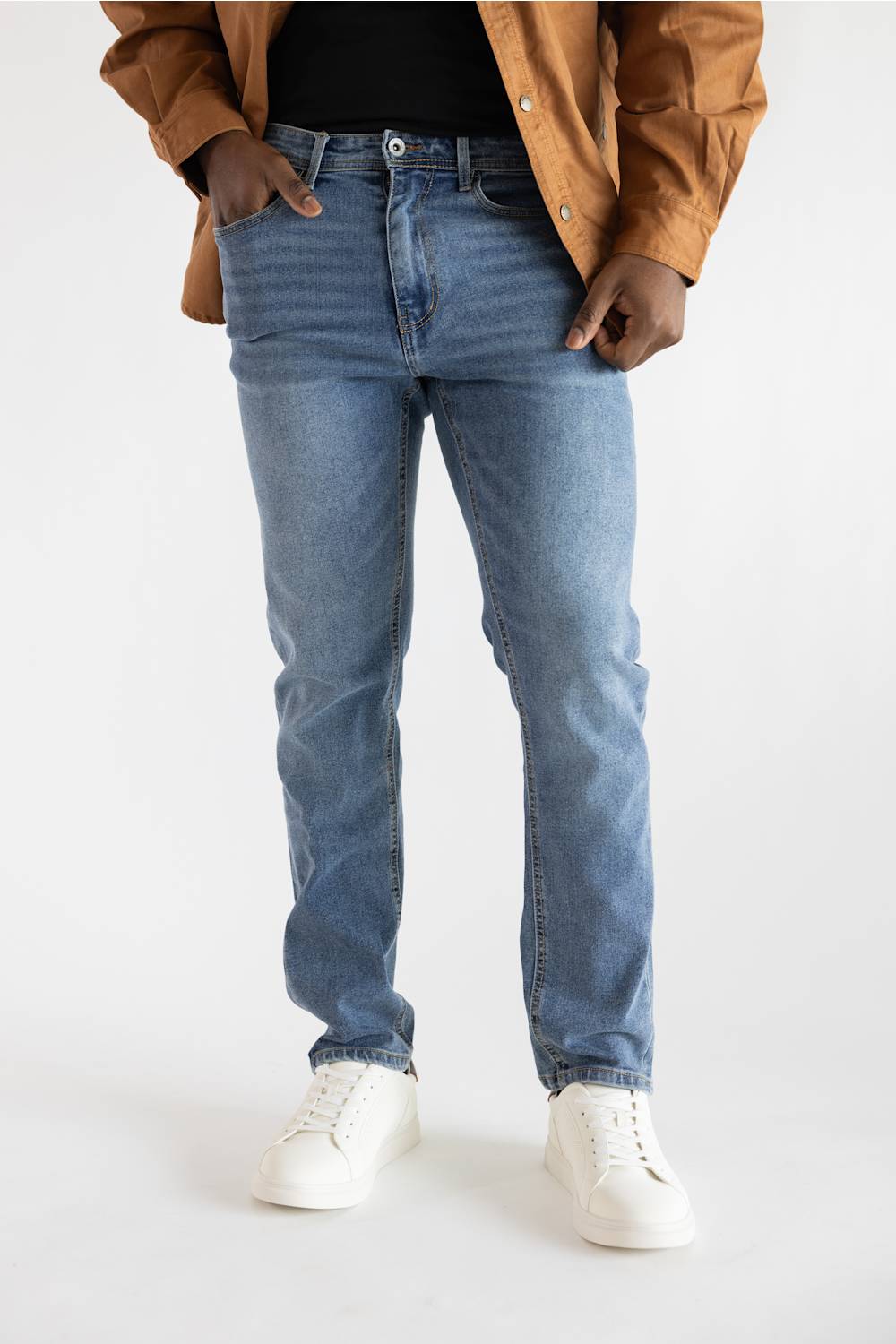 A person wearing light blue slim-fit jeans, styled with a black top, a brown jacket, and white sneakers, shown from the waist down against a plain white studio background.
