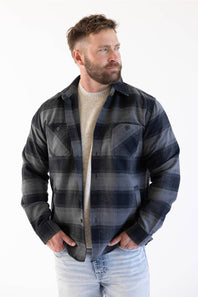 A man modeling the Weatherproof Vintage Lumberjack Plaid Shirt Jacket in "Phantom Night" (charcoal and dark navy plaid) worn open over a beige crewneck sweater and light-wash jeans with his hands in his pockets against a white studio background.