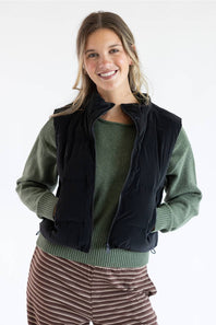 A smiling woman wearing a black sleeveless puffer vest over a sage green knit sweater and brown striped pants, photographed against a clean white studio background.