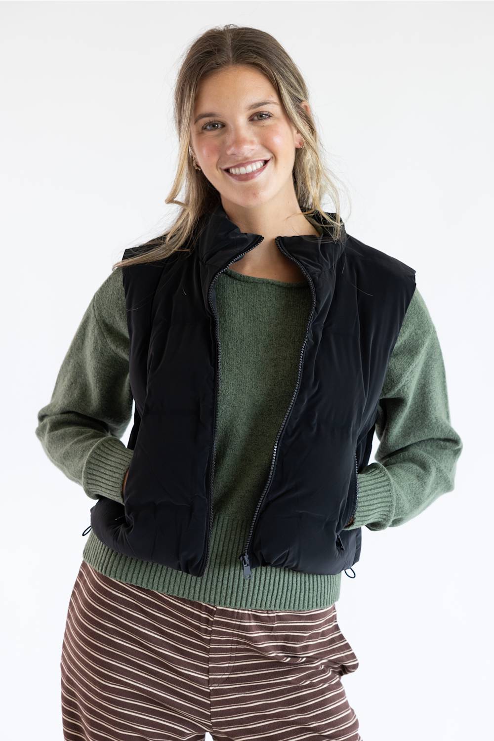A smiling woman wearing a black sleeveless puffer vest over a sage green knit sweater and brown striped pants, photographed against a clean white studio background.