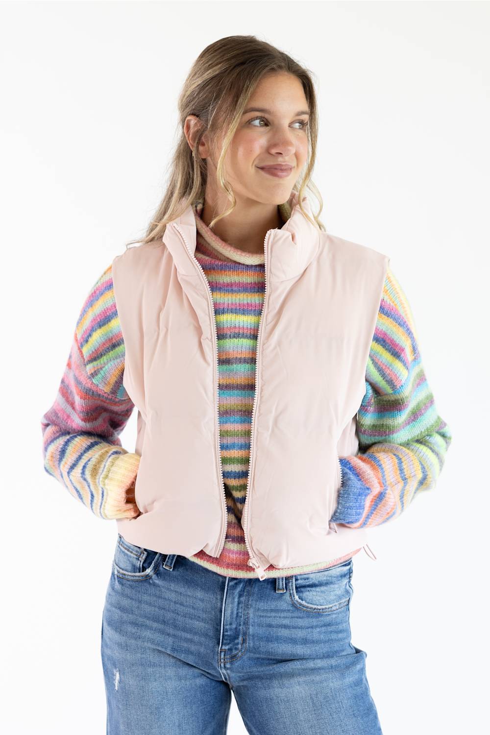 A woman wearing a pale pink puffer vest over a colorful striped knit sweater and blue jeans, photographed against a plain white studio background.