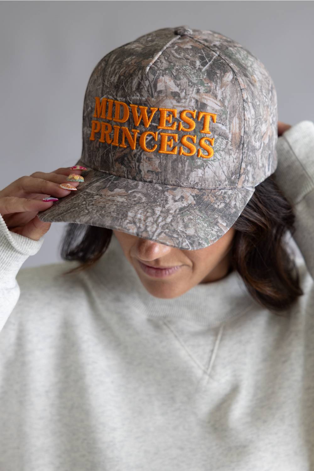 Camouflage cap with 'Midwest Princess' text worn by a woman in a gray sweatshirt.