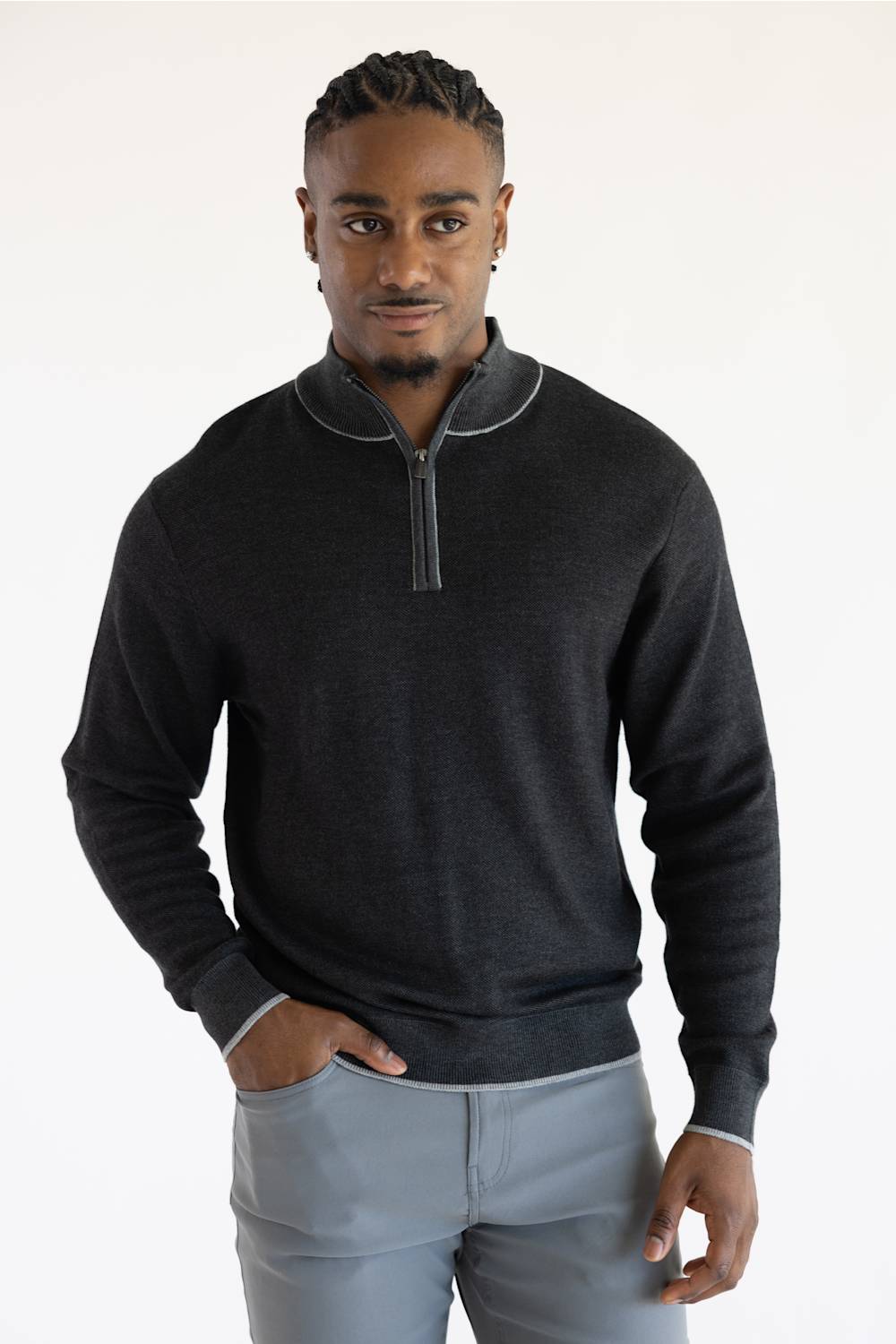 A man wearing a dark charcoal gray quarter-zip sweater with light gray piping details on the collar and cuffs, paired with light gray trousers, against a plain white studio background.