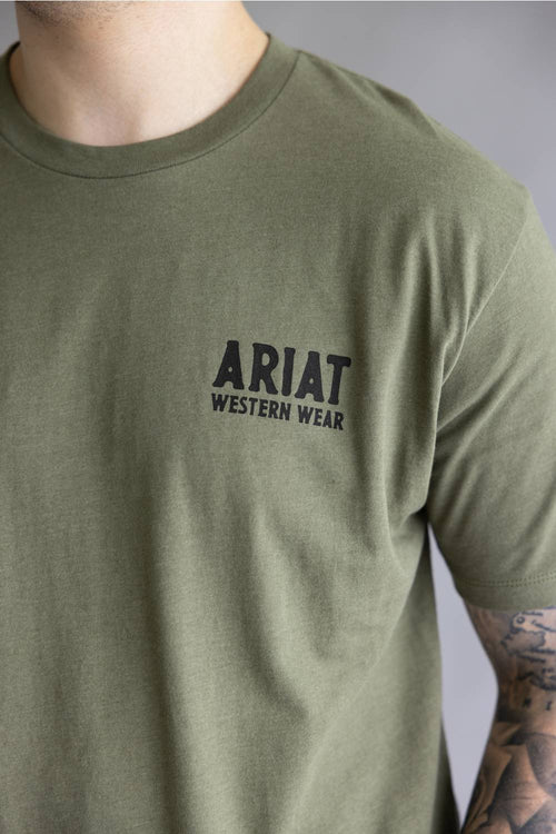 A close-up of a person wearing an olive green crewneck t-shirt with 'ARIAT WESTERN WEAR' printed in black on the left chest, set against a neutral gray background.