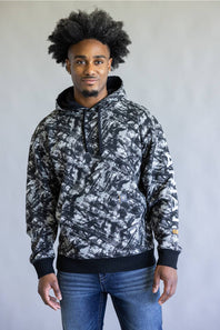 A man wearing a black and white abstract patterned hoodie with black ribbed cuffs, collar, and hem, paired with blue jeans, standing against a plain light gray wall.