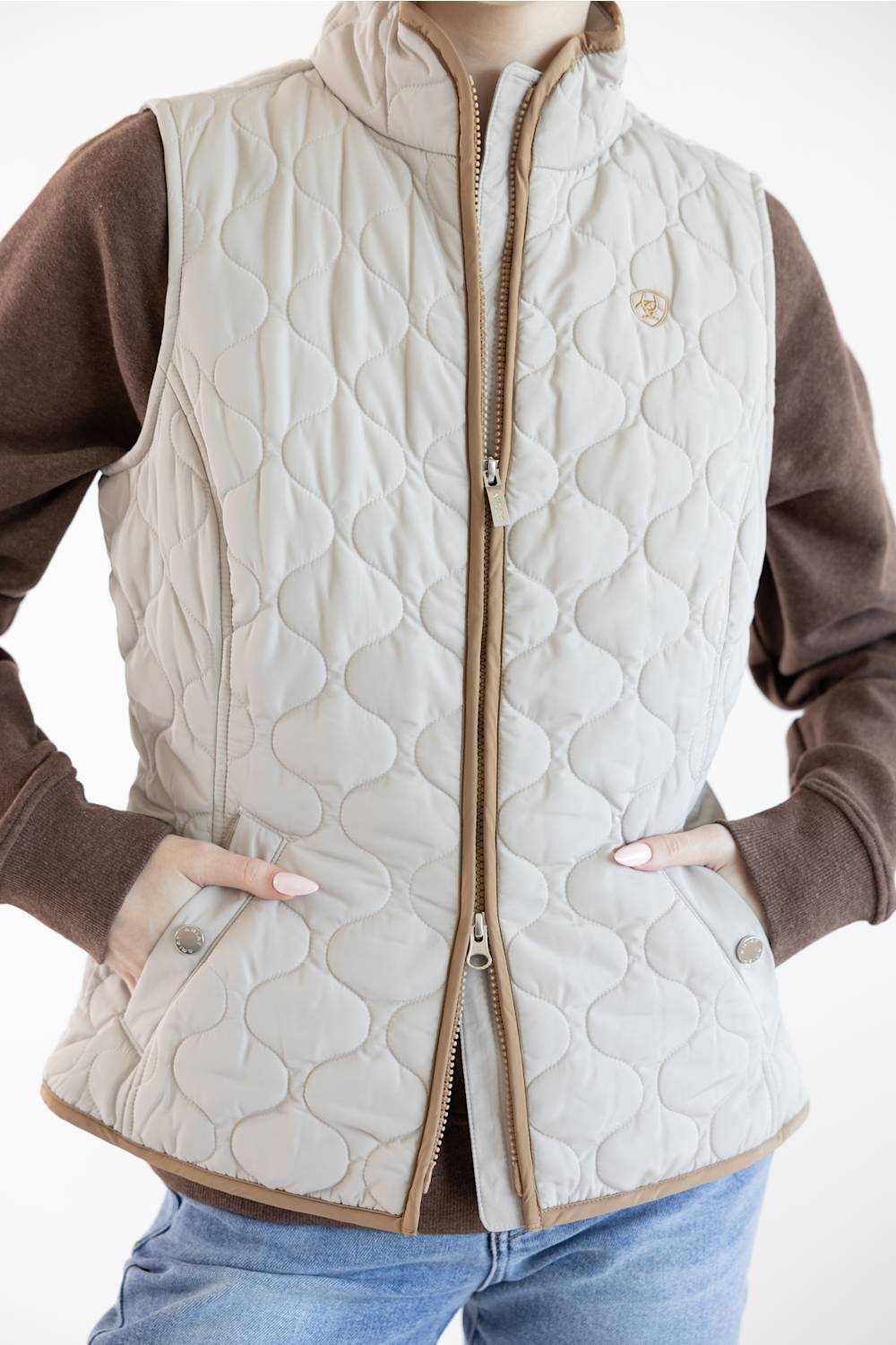 A quilted beige puffer vest with a brown zipper and trim, worn over a brown long-sleeve shirt and blue jeans.
