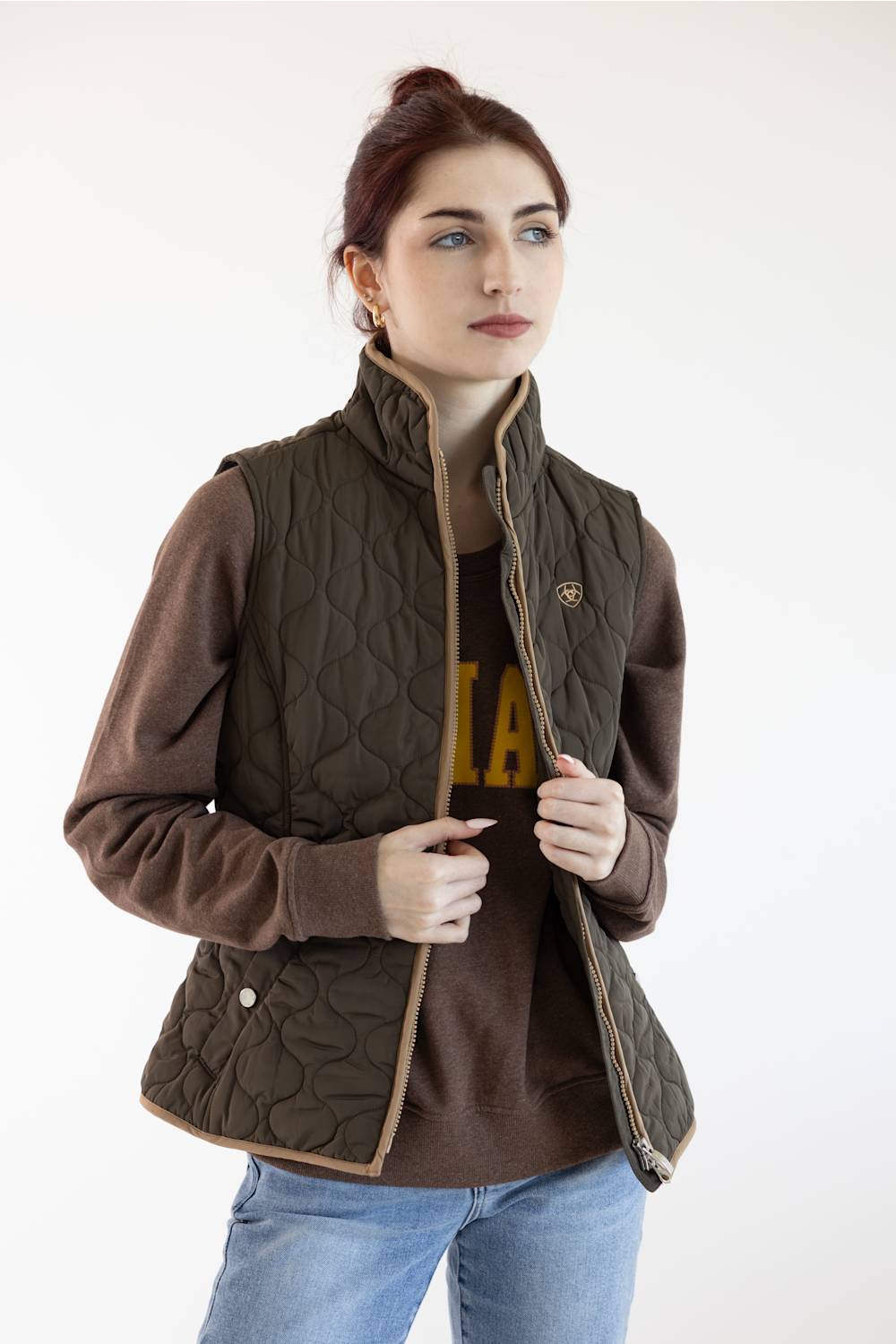 A woman wearing a brown quilted zip-front vest over a brown sweatshirt and blue jeans, photographed against a plain light background.