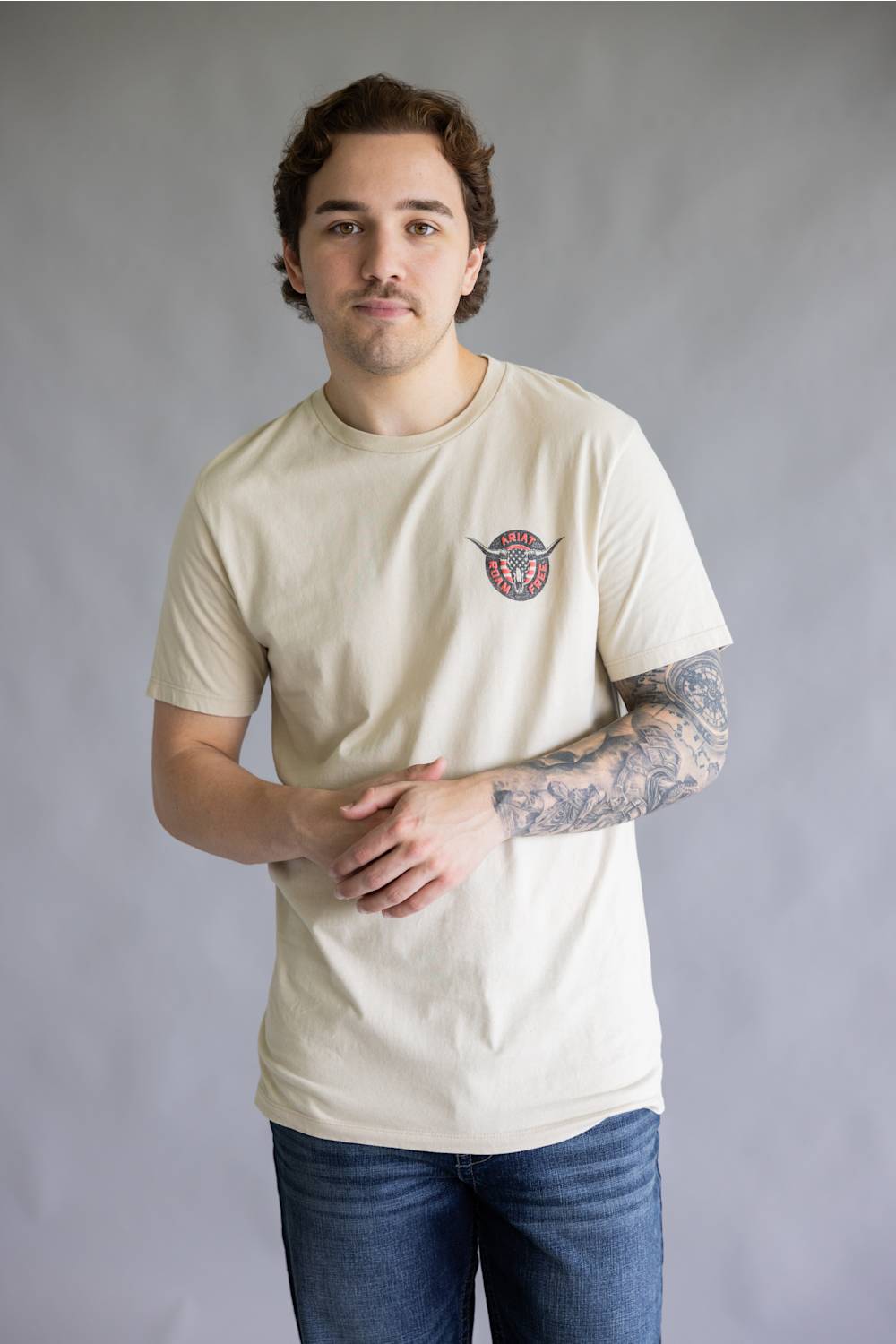 A man wearing a cream-colored crewneck t-shirt with a small circular graphic on the left chest, paired with blue jeans, standing against a neutral gray studio background.