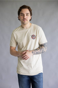 A man wearing a cream-colored crewneck t-shirt with a small circular graphic on the left chest, paired with blue jeans, standing against a neutral gray studio background.