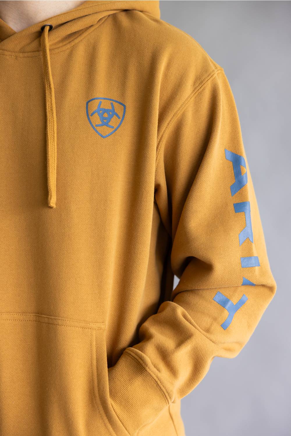 A close-up of a mustard yellow hoodie with a blue shield logo on the left chest and blue vertical lettering on the right sleeve, shown on a model against a gray background.
