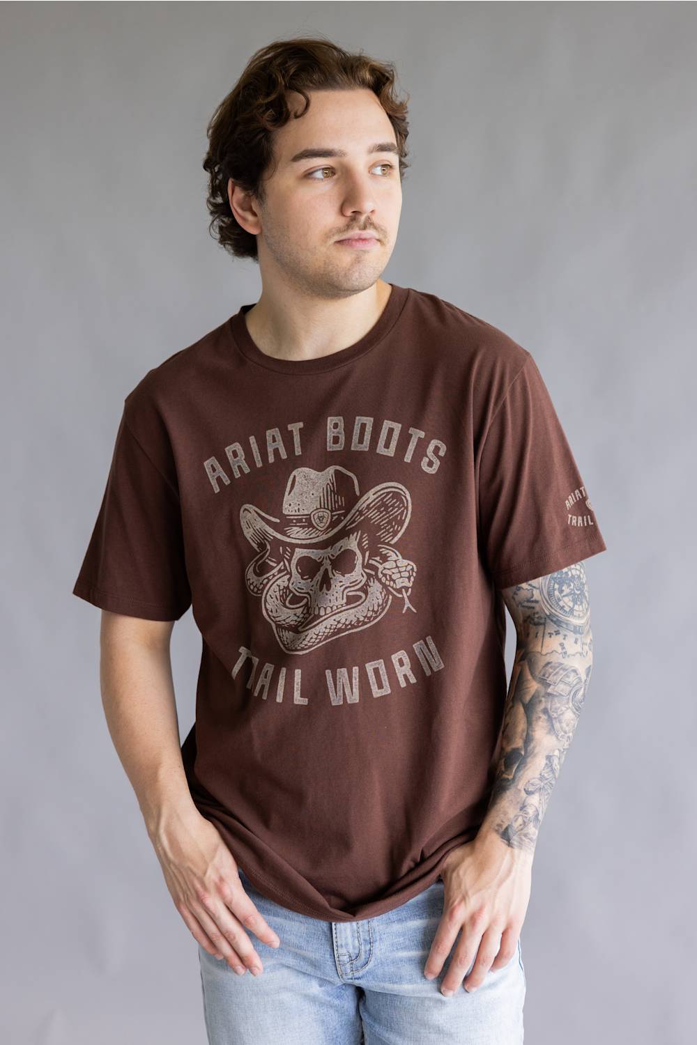 A man wearing a brown crewneck t-shirt with a distressed graphic of a cowboy hat and skull, featuring the text 'Ariat Boots Trail Worn'.